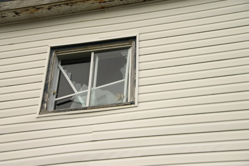 A wall with white siding with a broken window, shot from below
